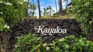 A stone wall with the name "Kanaloa" in cursive lettering, surrounded by lush greenery and palm trees.