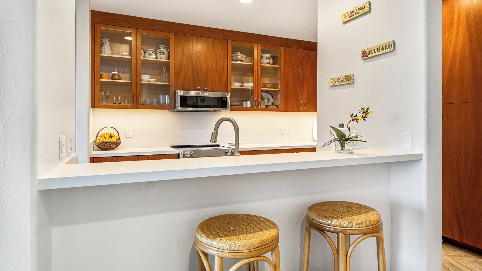 A kitchen with wooden cabinets, a stainless steel microwave, and a sink. There are two wicker stools and a small potted plant on the counter.
