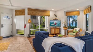 A spacious living room with a large blue sofa, a television, and a wicker chair. There are large windows with bamboo blinds, a fire extinguisher, and a door with a glass panel.