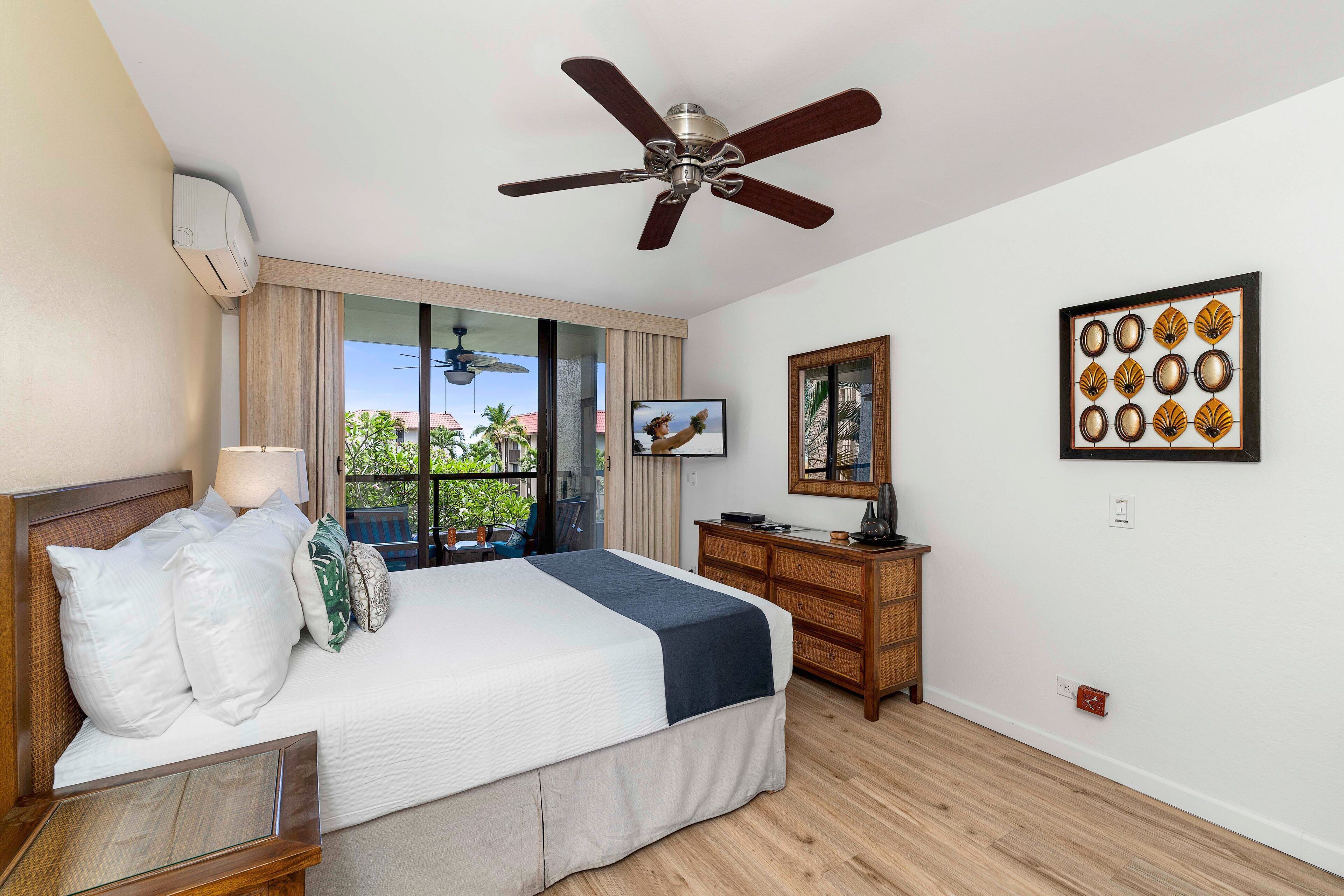 A hotel room with a bed, a wooden dresser, a ceiling fan, a TV, and a window with a view of palm trees.