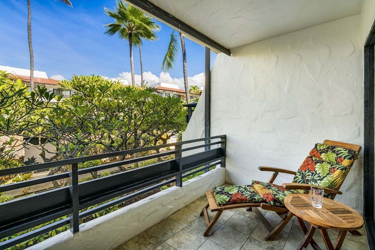 Un balcón con una silla y una mesa de madera, vistas a palmeras y un edificio al fondo.