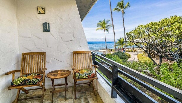 A balcony with two wooden chairs and a small table, overlooking a beach with palm trees and a clear view of the ocean. - Beautiful Ocean View - Updated - Wifi Included- Vrkcv (Kailua-Kona)