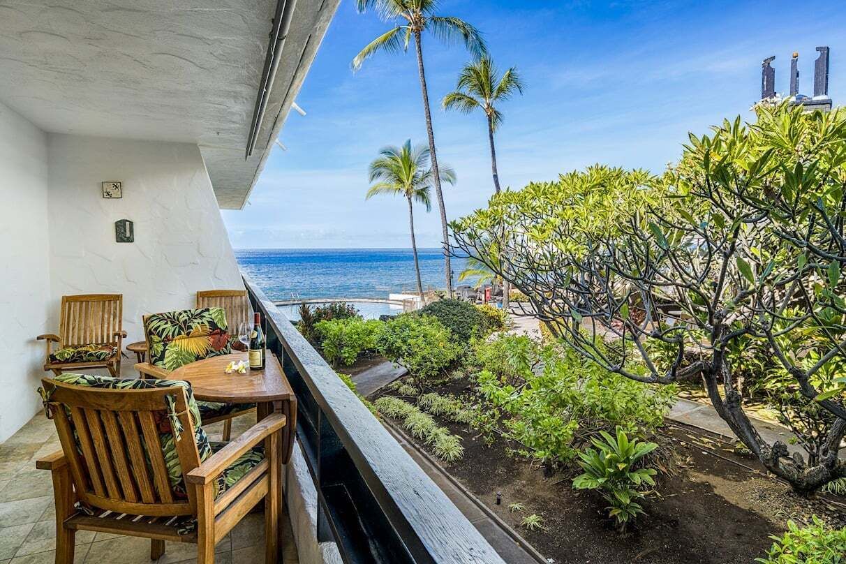 A balcony with wooden furniture, a view of the ocean, and tropical plants.