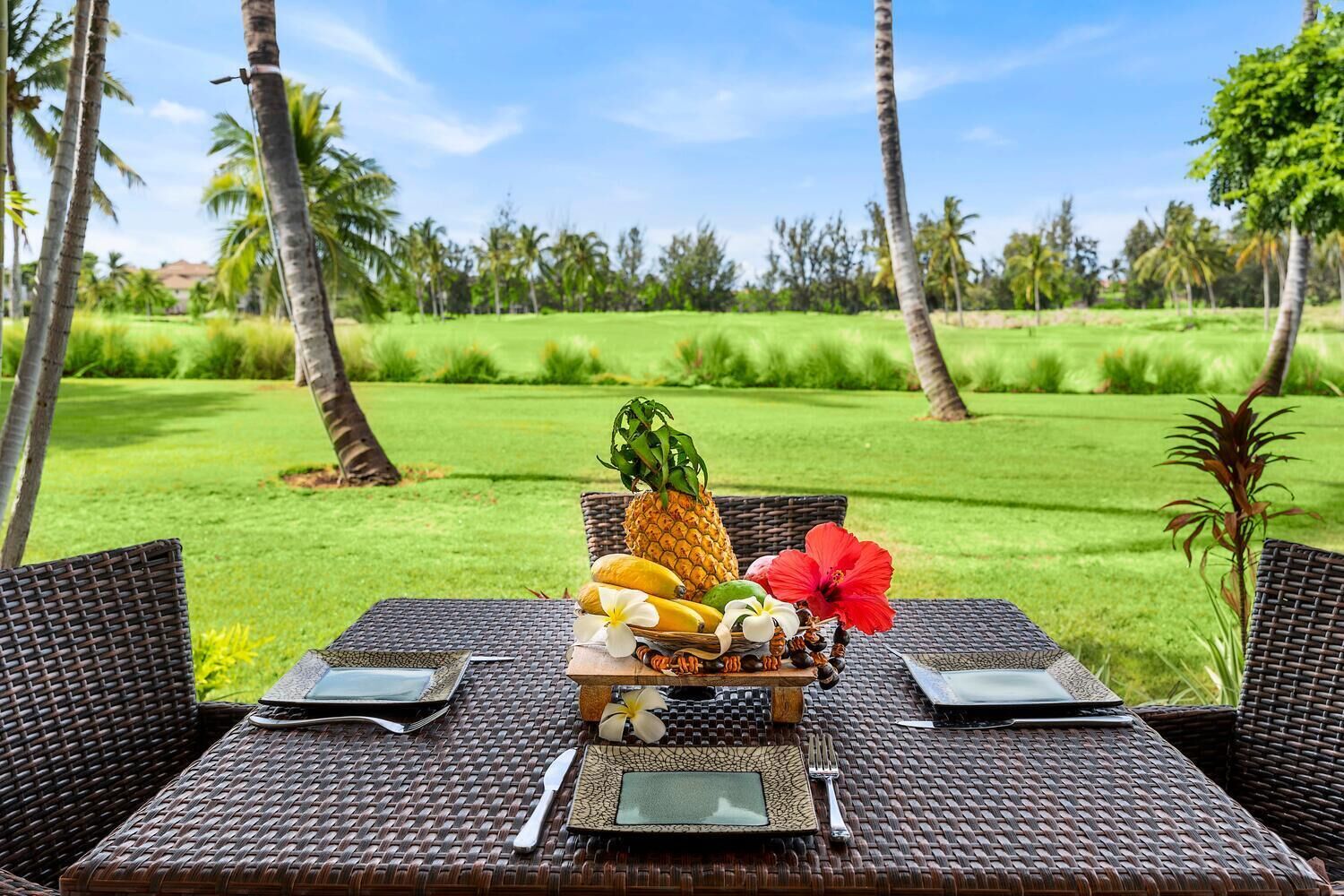 Une table dressée à l’extérieur, ornée d’un assortiment de fruits tropicaux, d’une fleur d’hibiscus rouge et d’une petite statue.