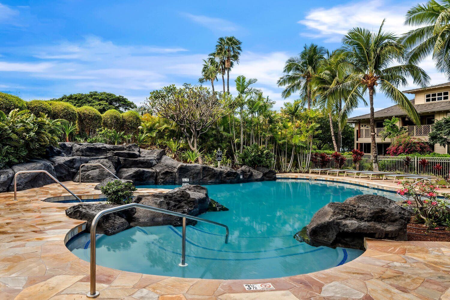 A pool with a rocky edge, surrounded by palm trees and a building with a balcony.