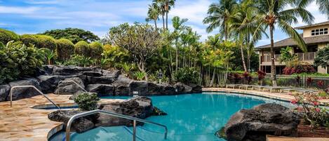 A pool with a rocky edge, surrounded by palm trees and a building with a balcony.