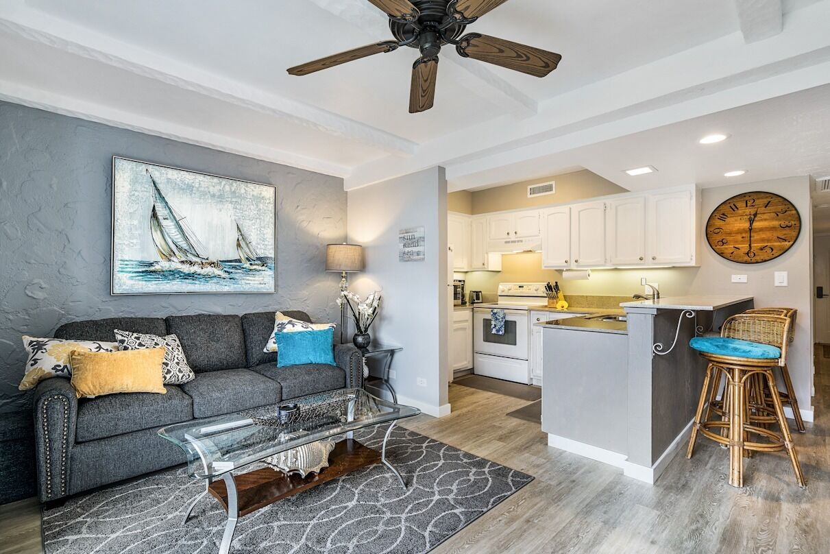 A modern living room with a gray sofa, a glass coffee table, and a painting of sailboats. A kitchen with white cabinets and a wooden island is visible in the background.