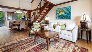 Amplia sala de estar con una mesa de centro de madera, un sofá blanco y una zona de comedor con sillas de mimbre. Una escalera con barandillas de madera conduce a un nivel superior.