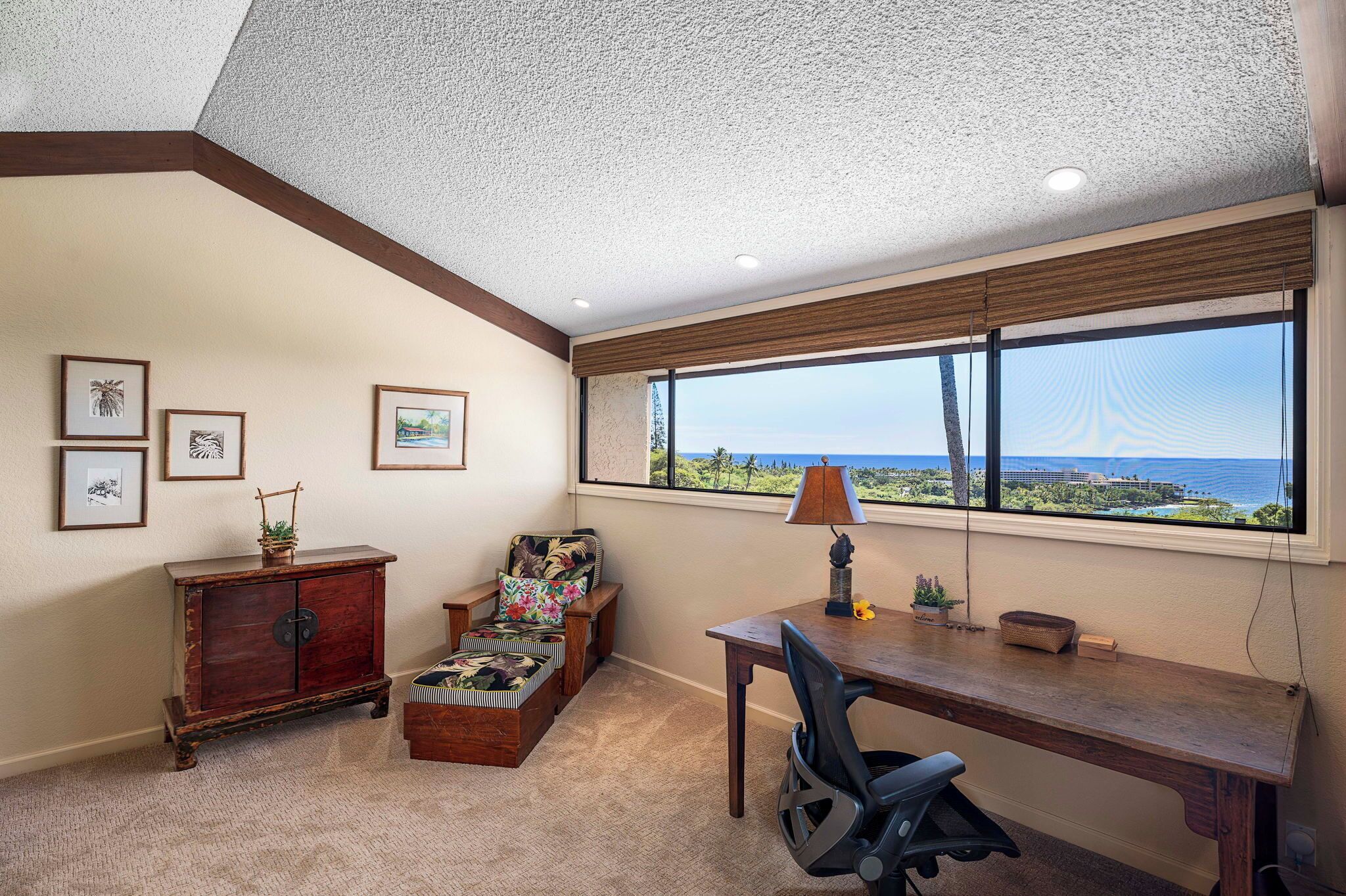 Una habitación con escritorio, silla y vistas al océano. Se ve una ventana con persianas, una lámpara y cuadros en la pared.