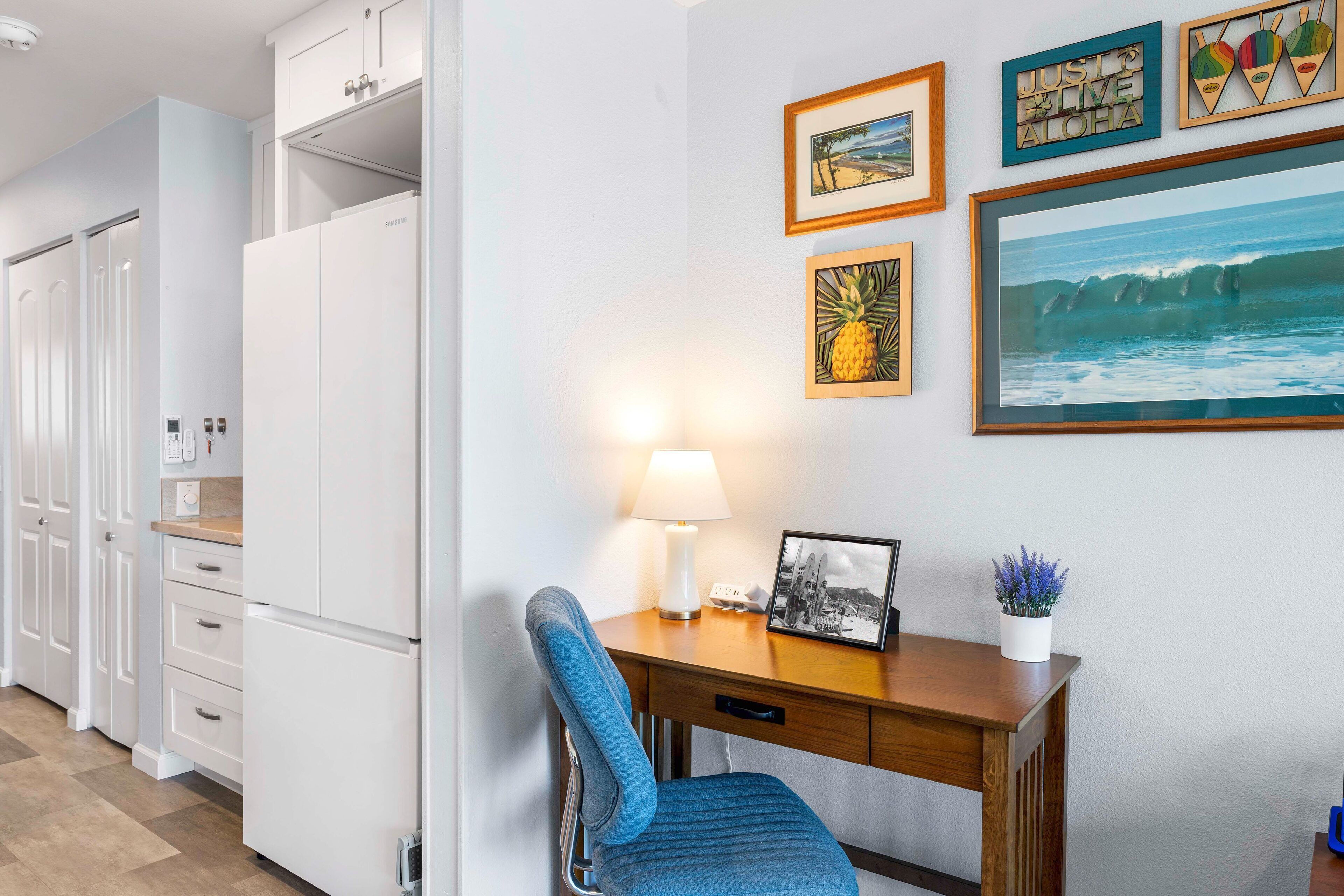 A kitchen with white cabinets, a wooden desk with a chair, and wall-mounted artwork including a framed picture of a beach and a framed text saying "JUST LIVE ALOHA".