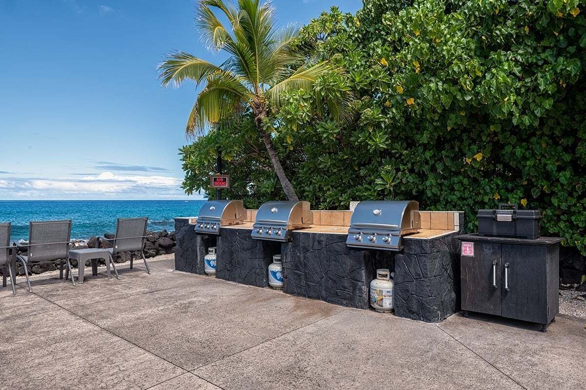 An outdoor grill area with multiple barbecue stations, a stone cabinet, and chairs near a beach.