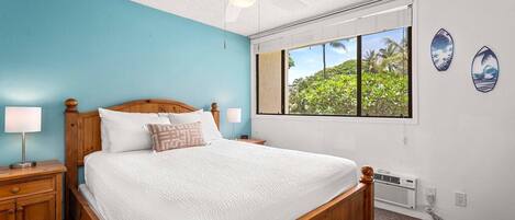 Un dormitorio con cama de madera, dos mesitas de noche con lámparas, un ventilador de techo, un ventanal con vistas a la vegetación y dos adornos de tablas de surf en la pared.