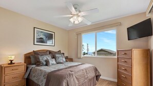 Un dormitorio con cama, mesita de noche, cómoda, ventilador de techo, televisor y ventana con vistas a edificios.