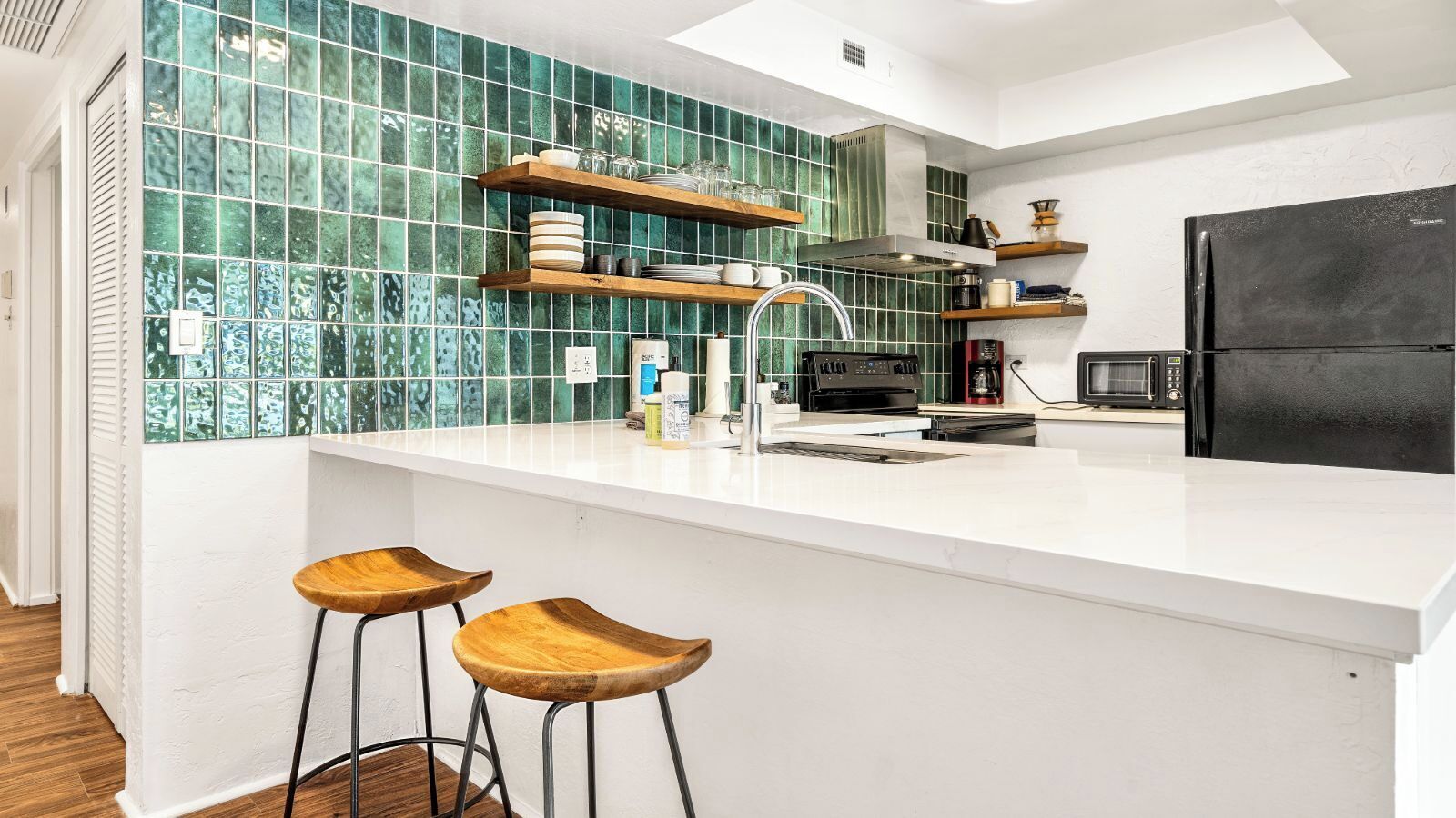 Una cocina moderna con encimera blanca, salpicadero de azulejos verdes, taburetes de madera y un refrigerador negro.
