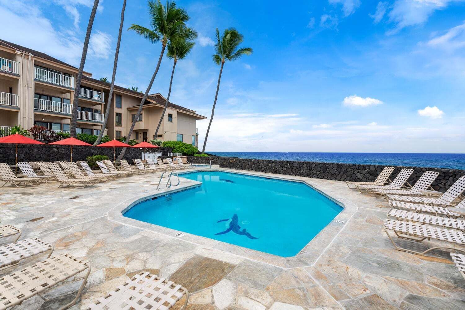 A pool with lounge chairs and umbrellas, a stone deck, and a building with balconies.