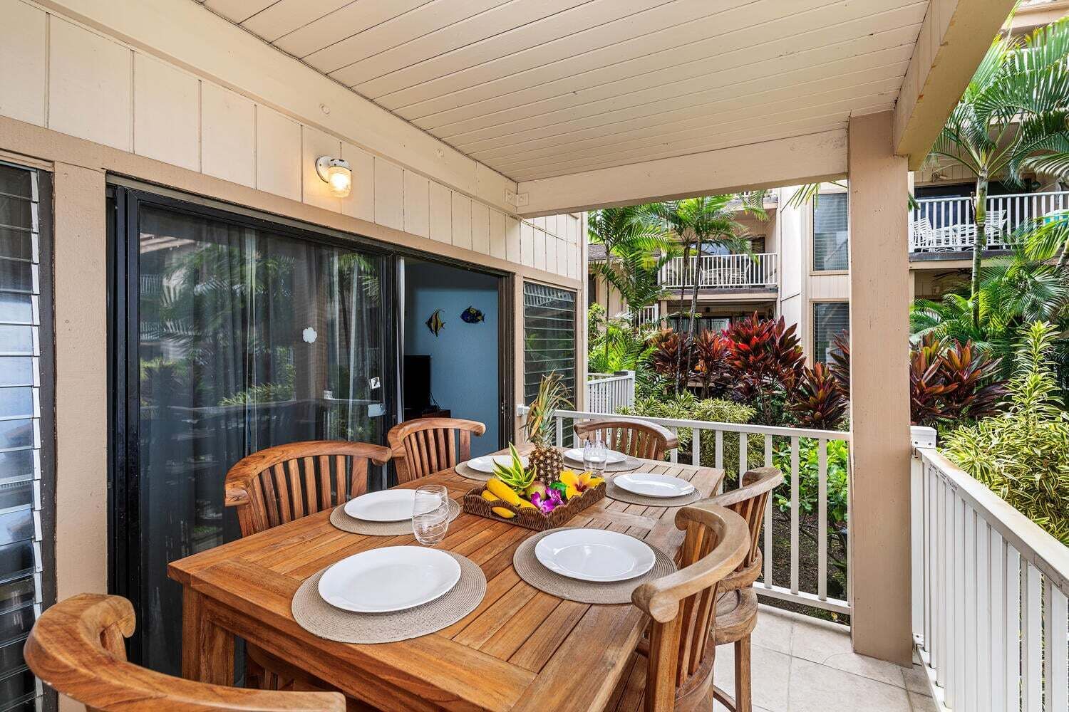 A wooden table set for four with plates, glasses, and a fruit basket, surrounded by chairs, on a balcony with a view of a lush garden and a building in the background.