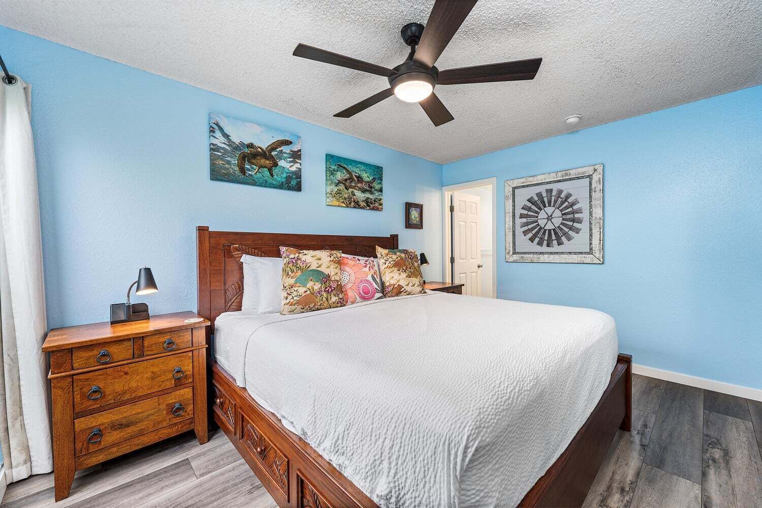 A bedroom with a bed, wooden nightstand, ceiling fan, and artwork on the walls.