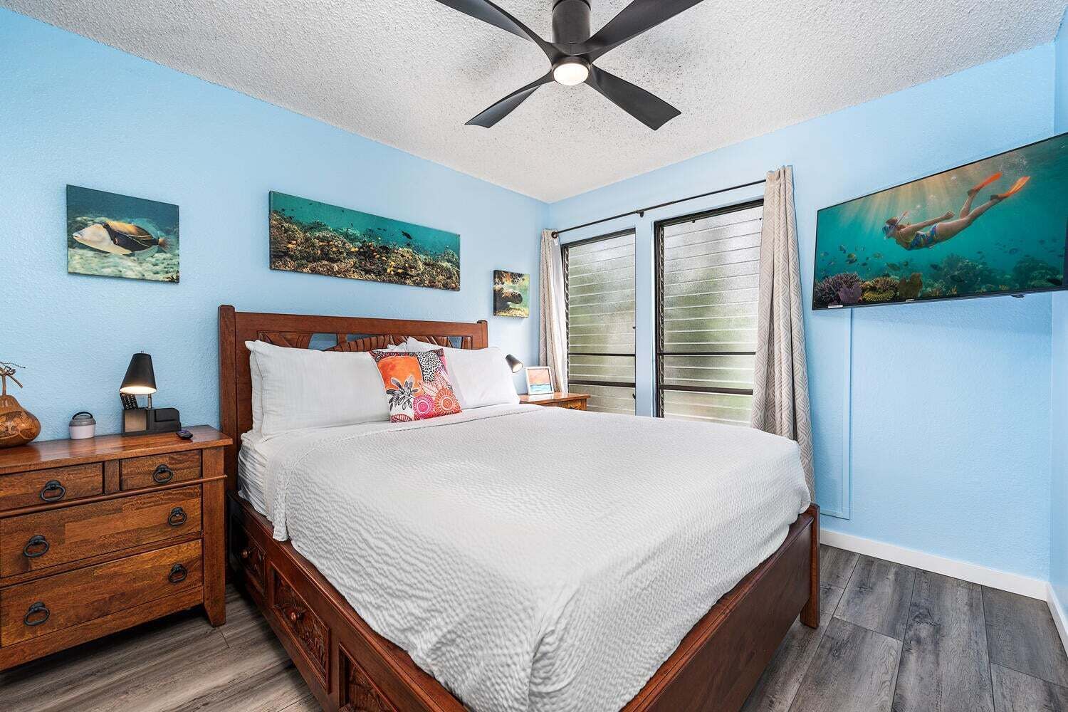 A bedroom with a bed, wooden nightstand, dresser, ceiling fan, and artwork.