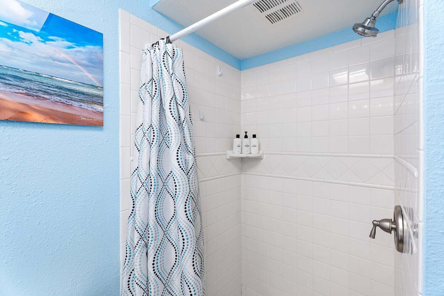 Un baño con paredes de azulejos blancos, una ducha con cabezal de ducha tipo lluvia y una cortina de ducha con diseño geométrico.
