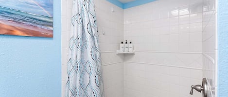 Un baño con paredes de azulejos blancos, una ducha con cabezal de ducha tipo lluvia y una cortina de ducha con diseño geométrico.
