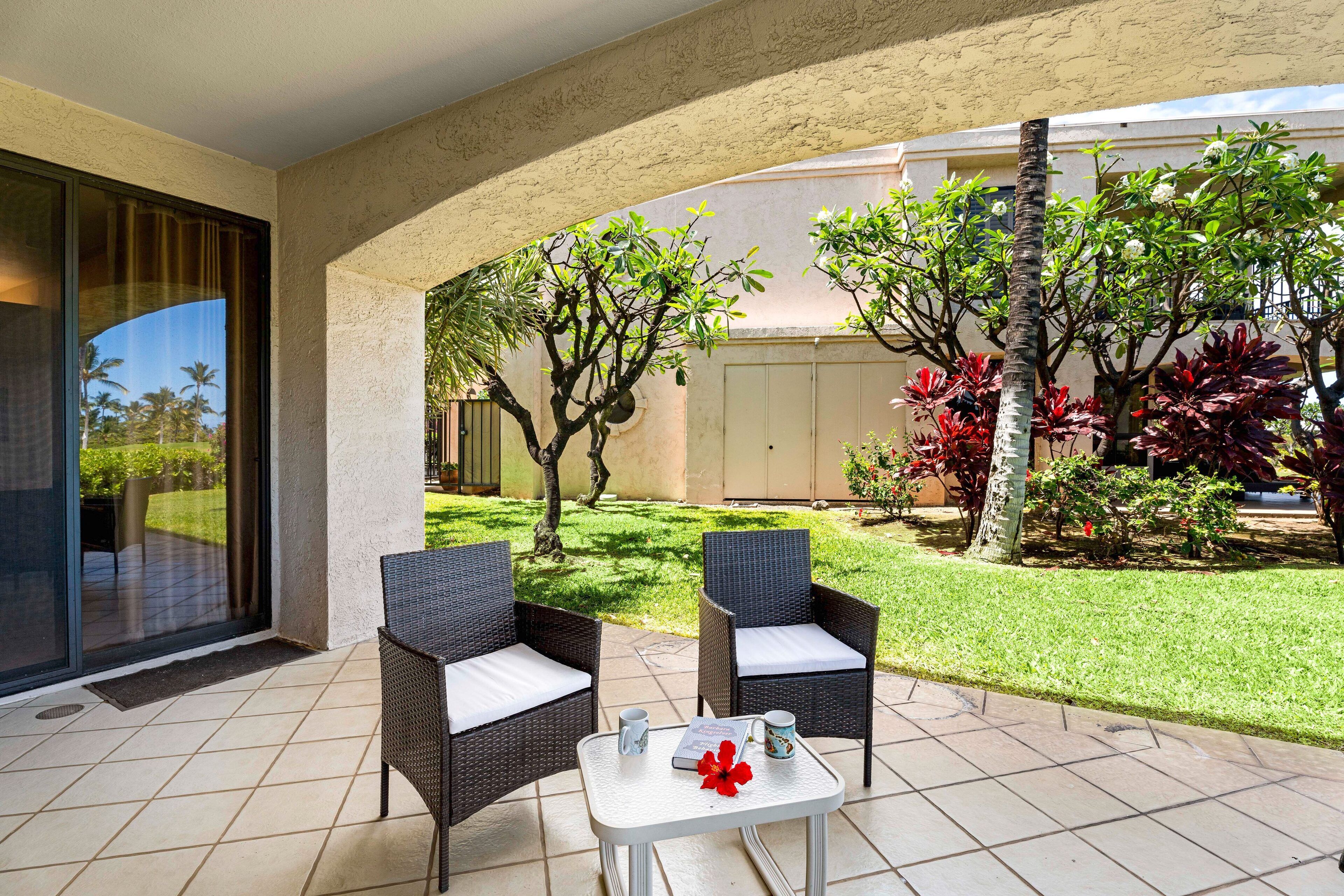 A patio with two wicker chairs and a small table, surrounded by a garden with various plants and a view of a building in the background.
