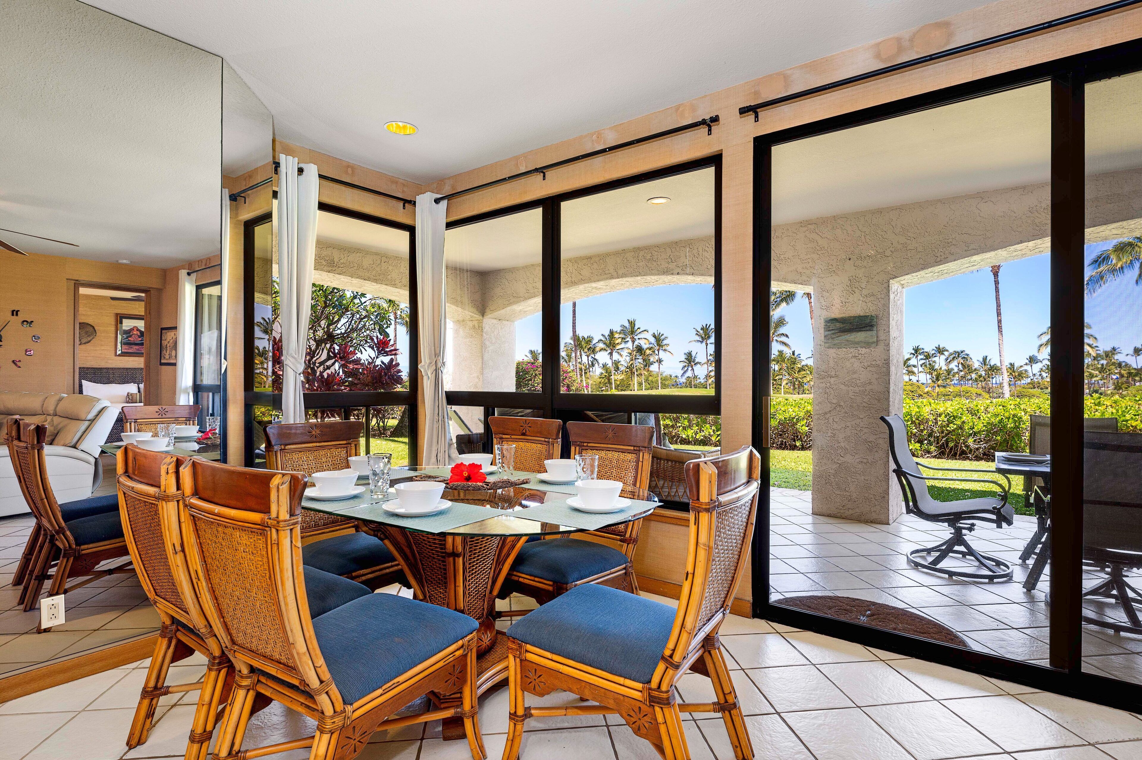 Un coin repas avec une table ronde, des chaises en osier et une vue sur les palmiers, visible à travers de grandes portes vitrées.