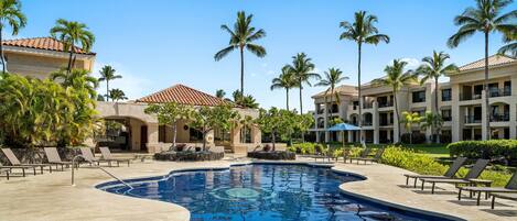 A resort pool area with lounge chairs, palm trees, and a building with a red-tiled roof.