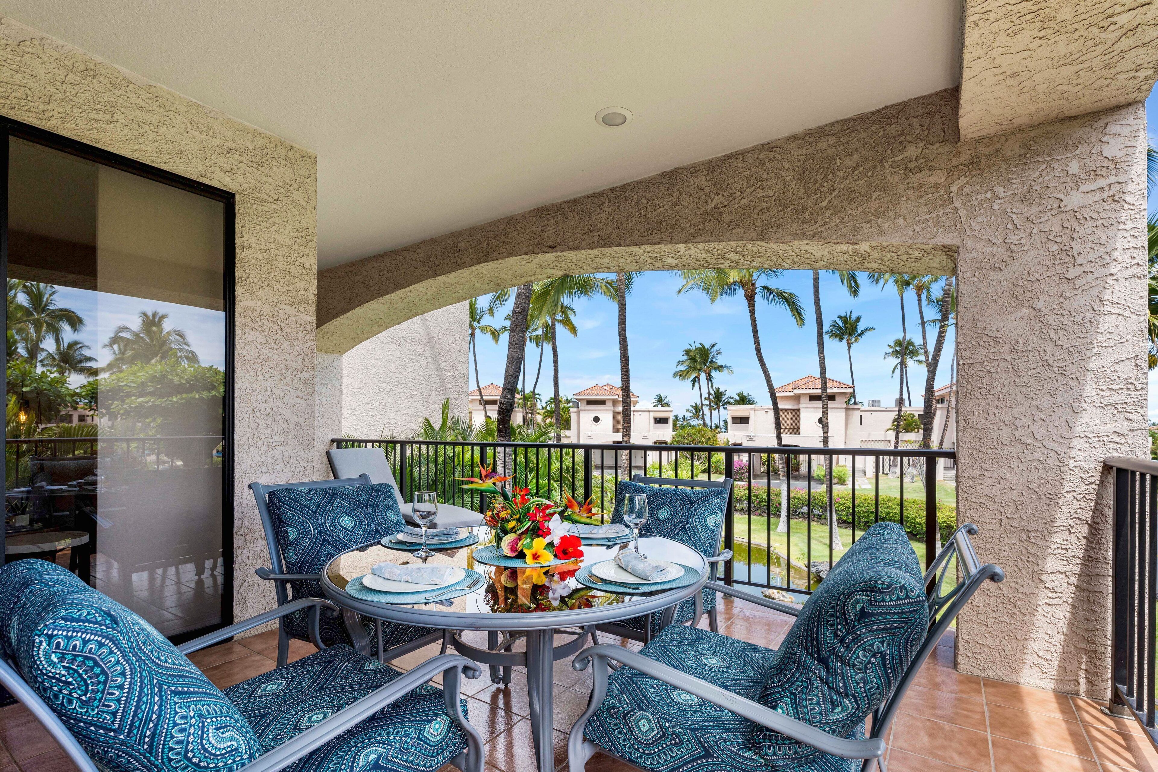 A balcony with a table set for dining, featuring patterned cushions and a floral centerpiece.