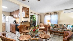 A compact kitchen with wooden cabinets, a white refrigerator, and a dining area with a glass table surrounded by wicker placemats and wooden chairs.