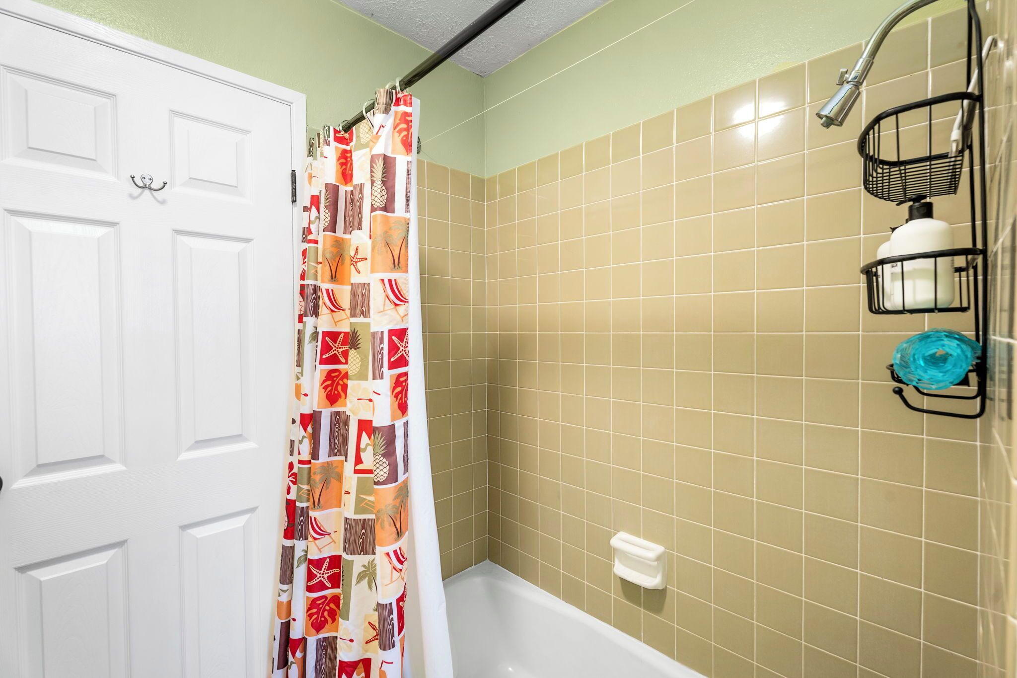 Un baño con azulejos color beige, una cortina de ducha con motivos tropicales, una bañera blanca y un perchero con una toalla azul.