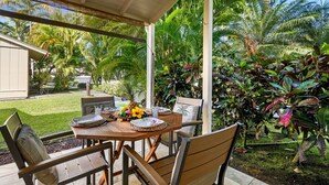 An outdoor patio with a wooden table set for dining, surrounded by chairs, and a view of a lush garden with various plants and a palm tree.