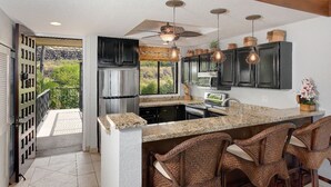 A kitchen with a granite countertop, dark cabinets, and a built-in island with bar stools. - Upgraded 2BD Plus Loft - Amazing Golf & Ocean Views - In-unit AC - Vrkcv (Kailua-Kona)
