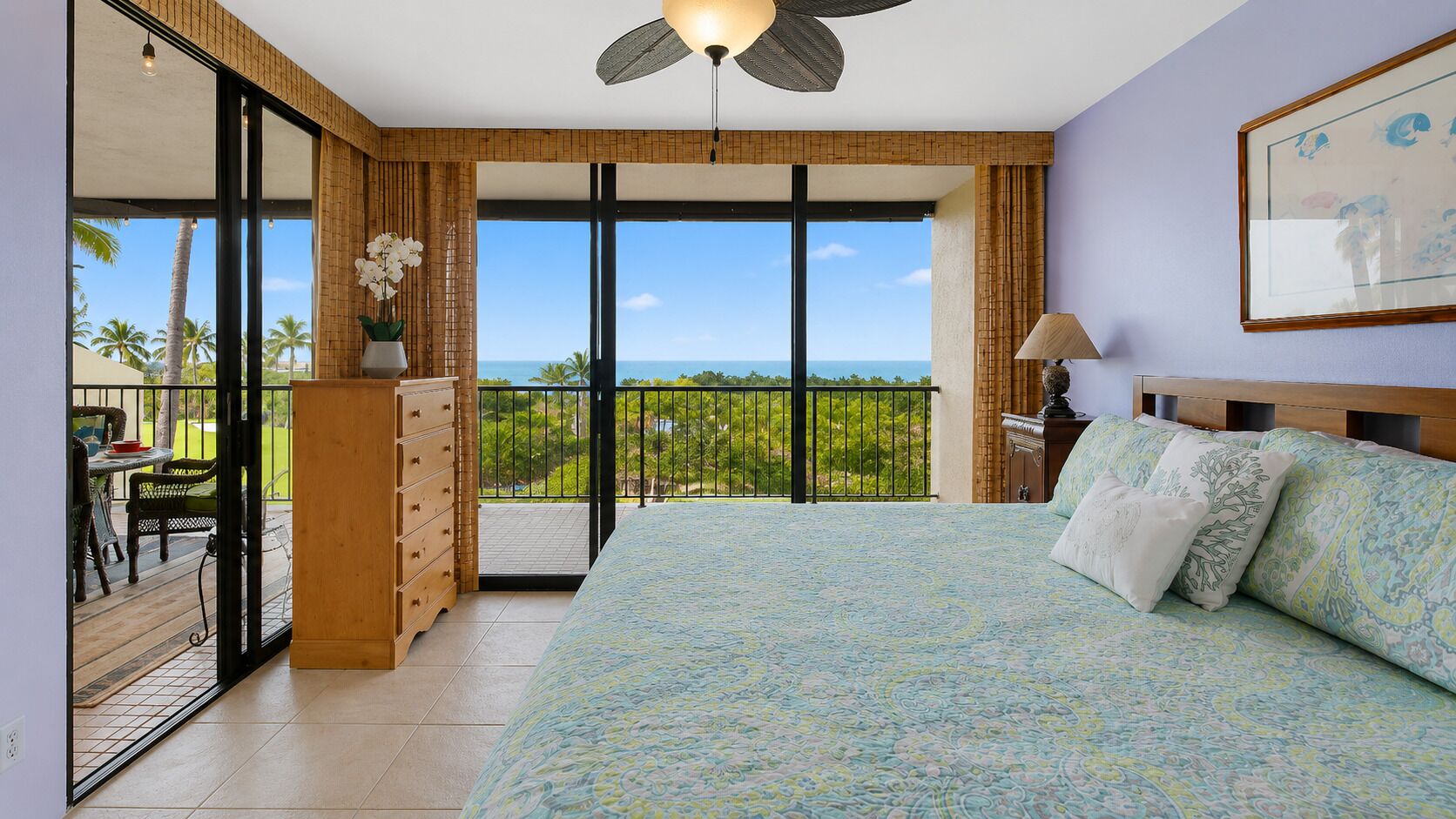 Une chambre avec un grand lit, une commode en bois, un balcon avec vue et un ventilateur de plafond.