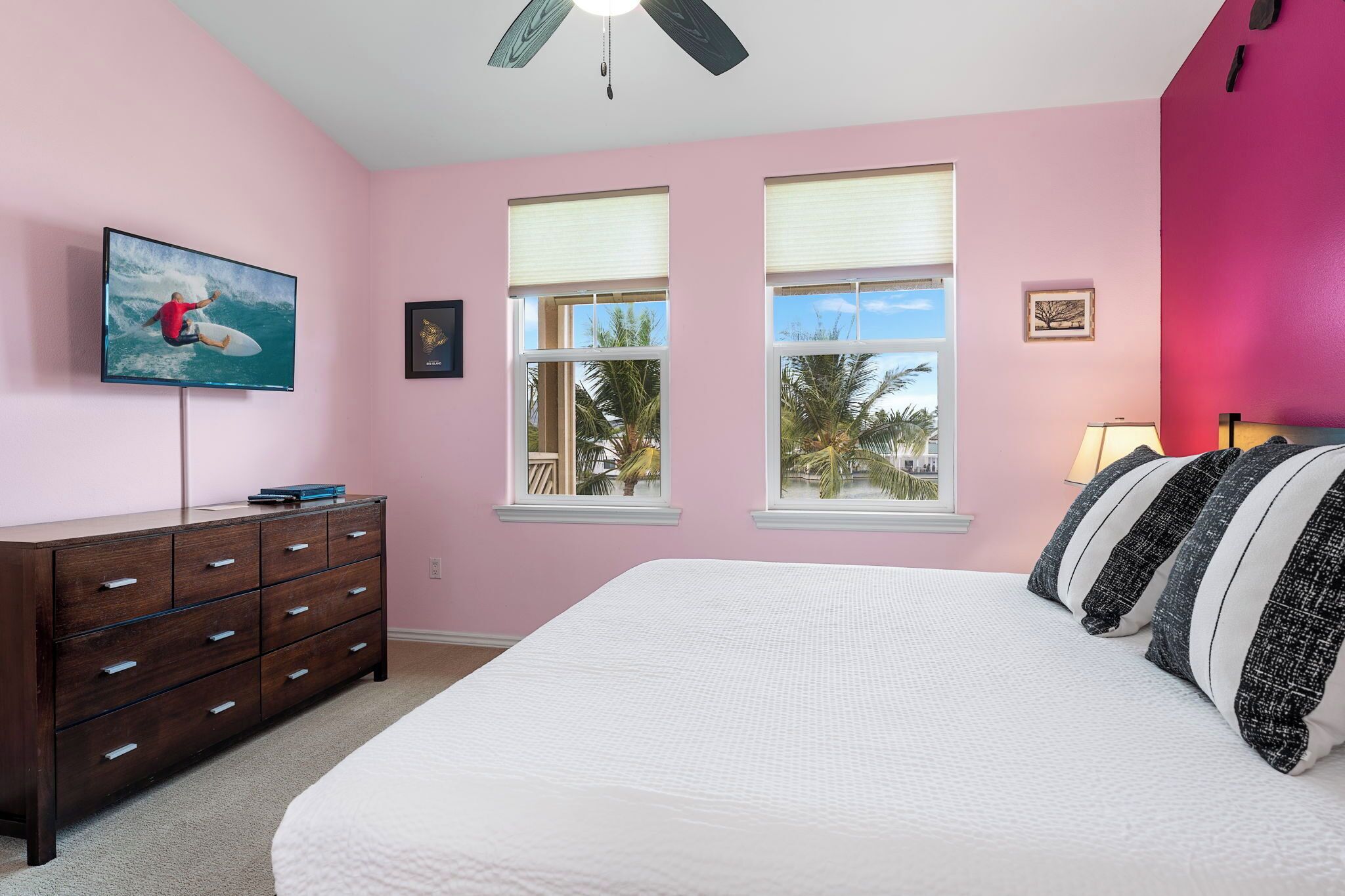 A bedroom with pink walls, a large bed with white bedding, a dark wooden dresser, a flat-screen TV on the wall, and two windows with blinds.