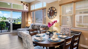 A dining area with a round table set for four, a sofa, and a large wall clock. - Concierge Included - Ocean View - Walk to Beach - Central AC - Vrkcv (Kailua-Kona)