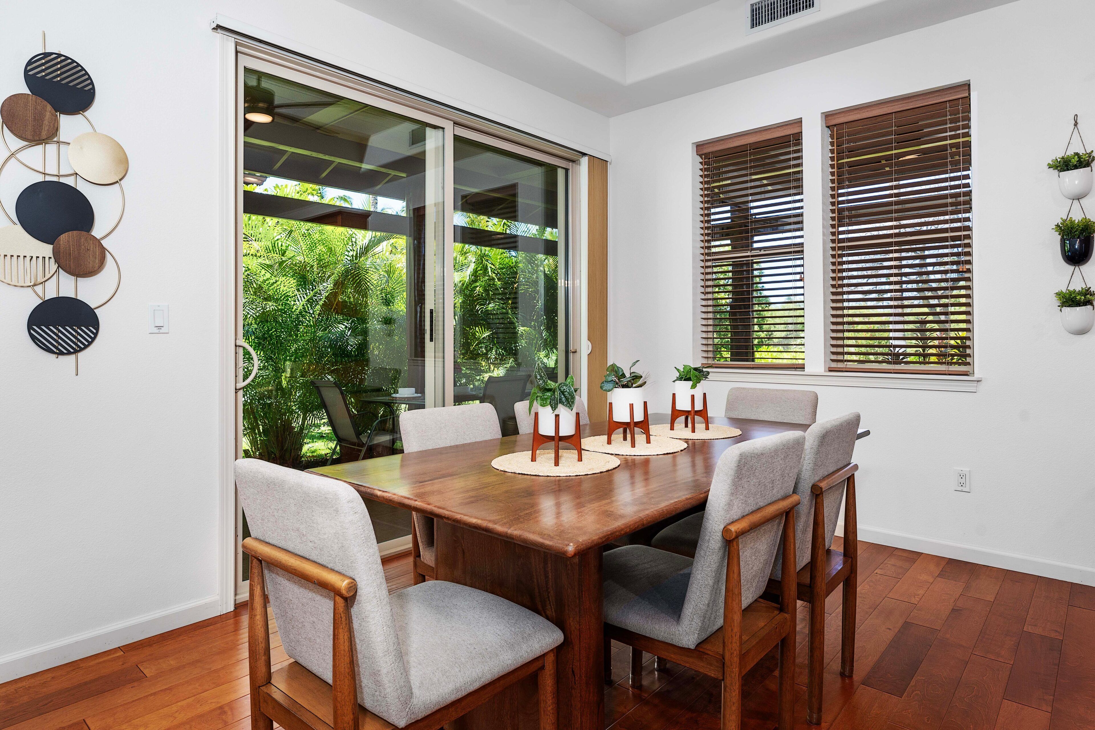 Un coin repas avec une table en bois et six chaises, de grandes fenêtres équipées de stores et une vue sur la verdure à l’extérieur.