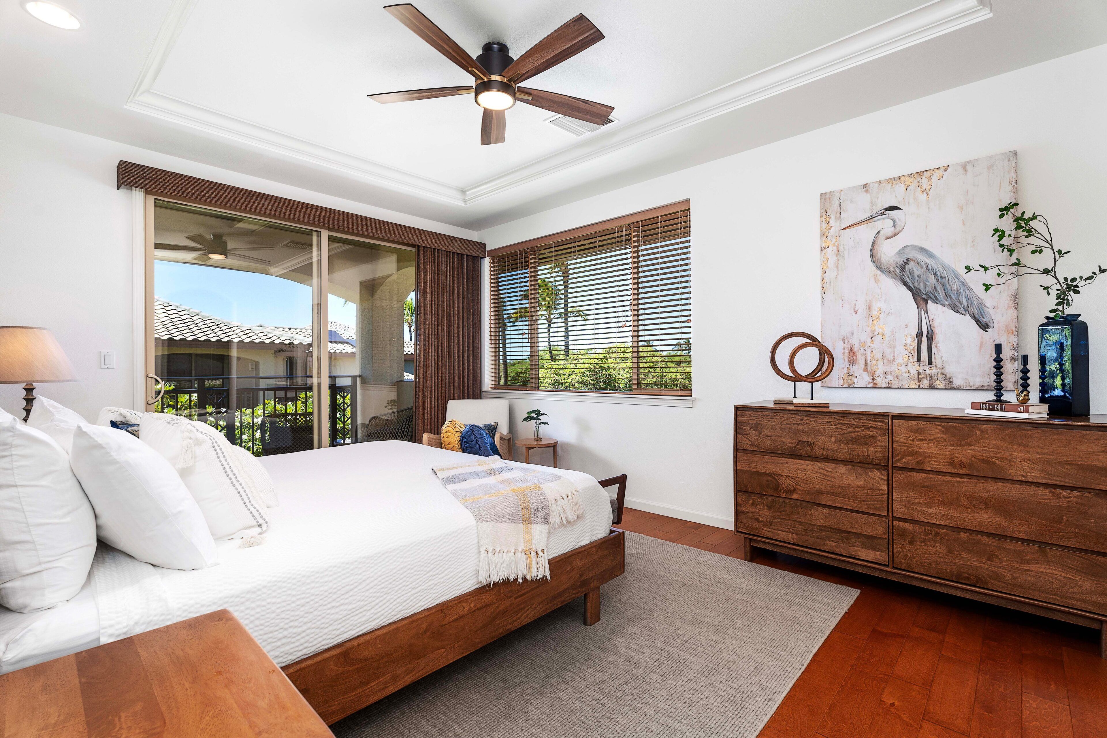 A bedroom with a large bed, wooden headboard, a ceiling fan, a window with blinds, a wooden dresser, and a painting of a bird.