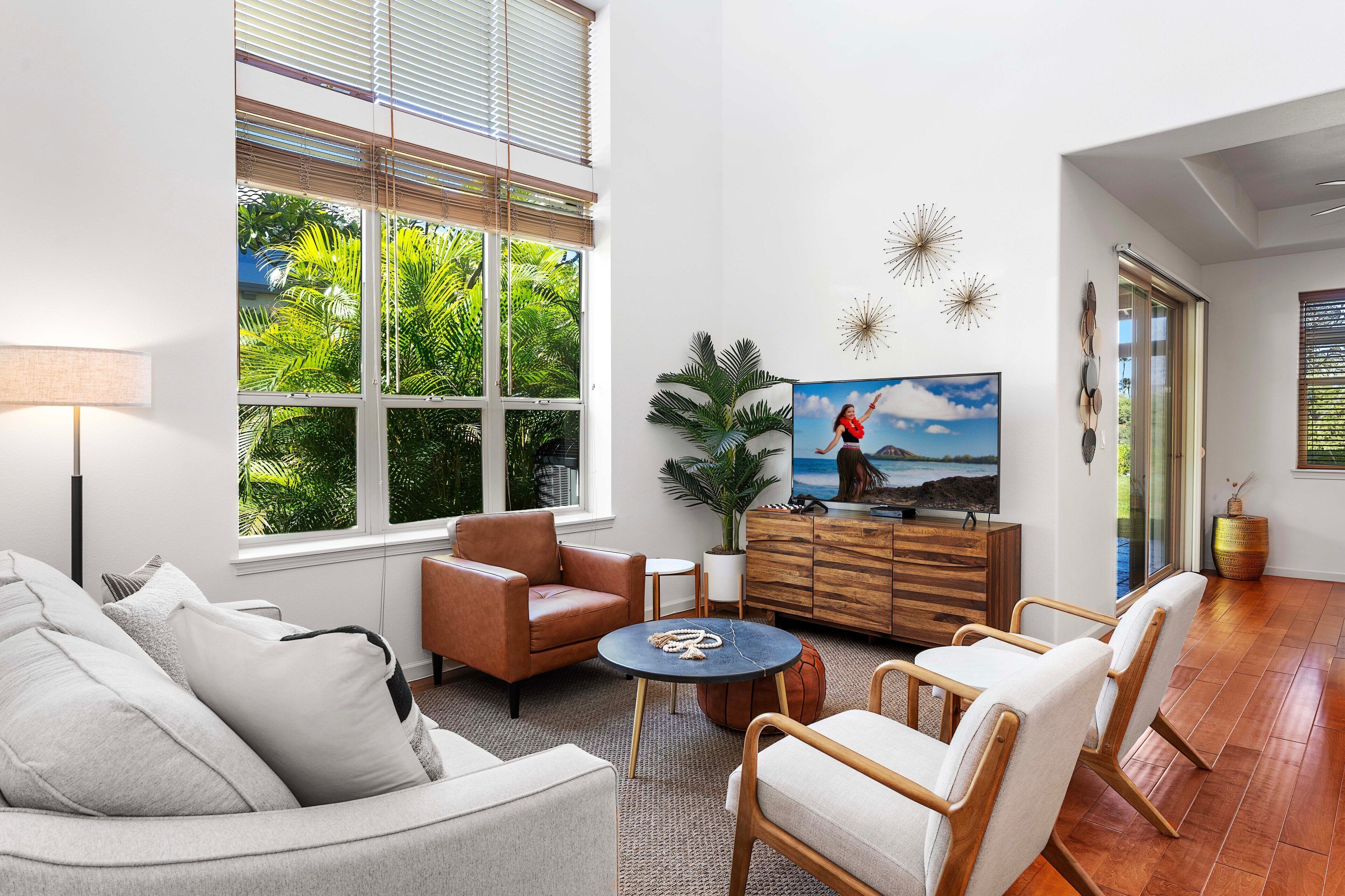 Un salon moderne avec un canapé, des fauteuils, une table basse et un téléviseur. On y trouve une grande fenêtre avec des stores, une plante en pot et un meuble en bois.