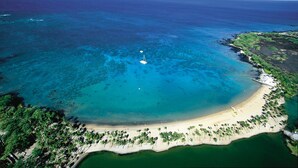 Vue aérienne d’une plage aux eaux turquoise et limpides, avec un rivage sablonneux et un petit voilier ancré près du rivage.