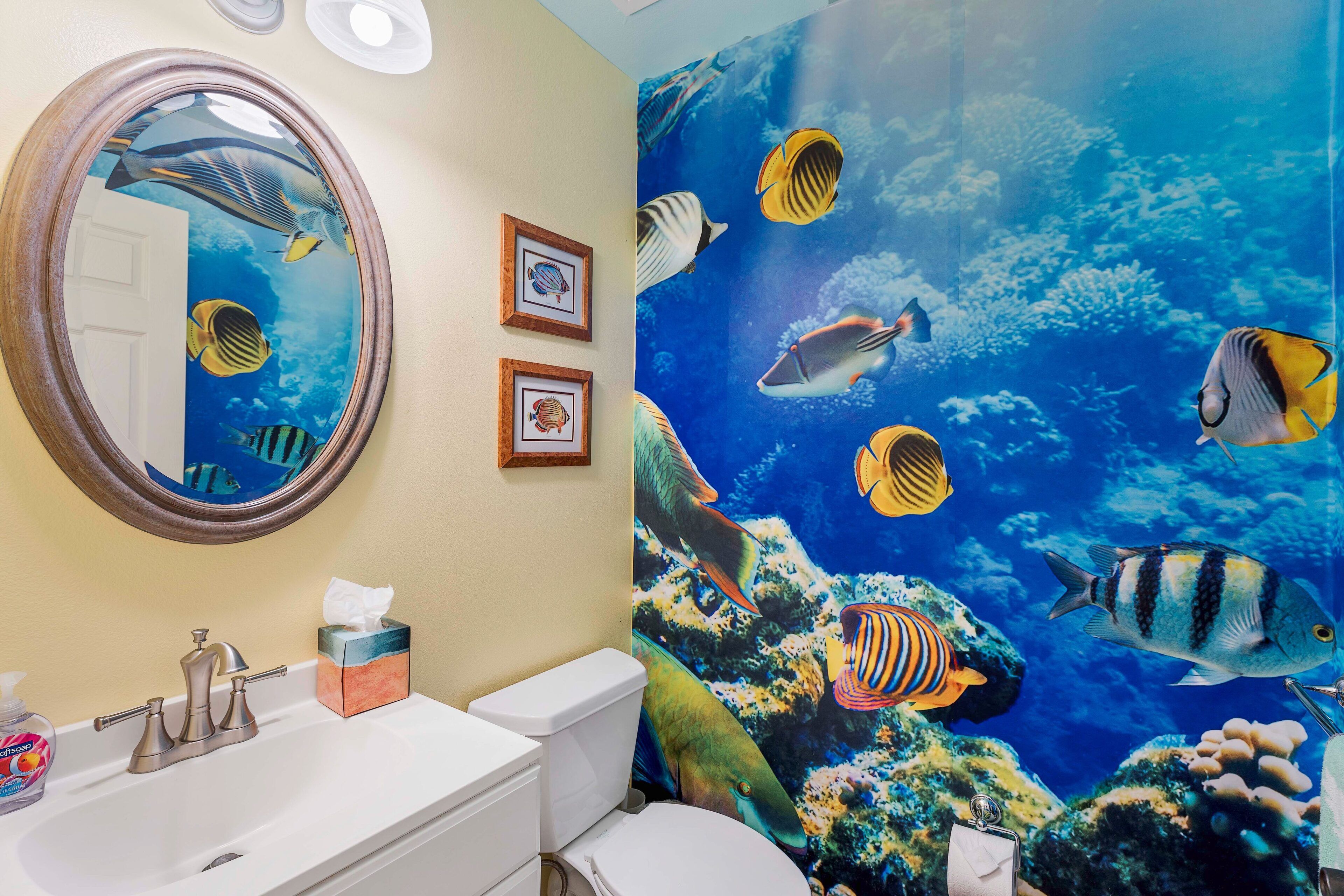 A bathroom with a large mirror, a sink, and a toilet. There is a framed picture on the wall and a tissue box on the sink.