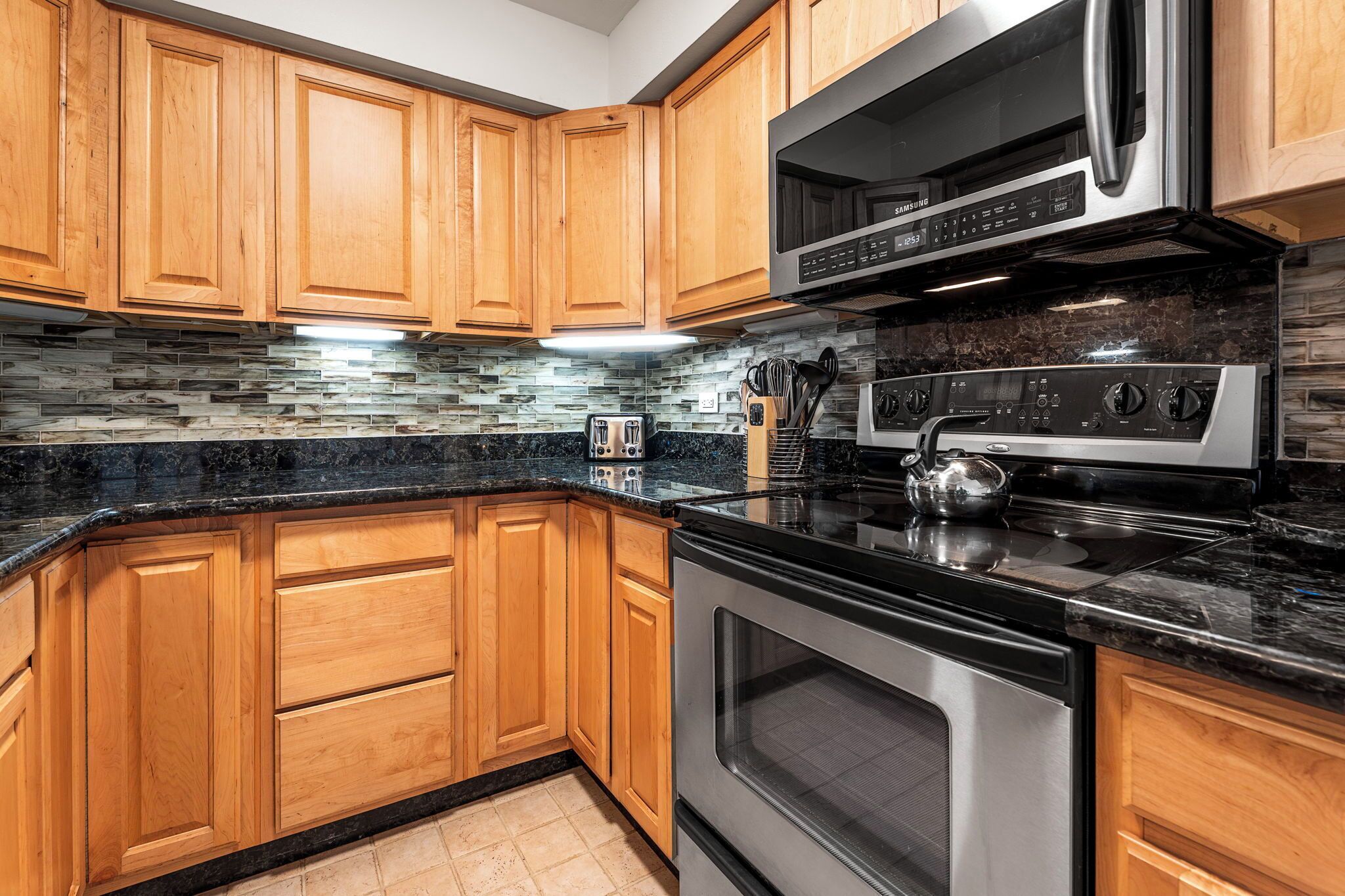 A kitchen with wooden cabinets, a black countertop, a stainless steel microwave, and a built-in oven.