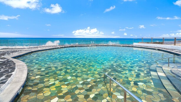 Une piscine aux eaux cristallines, dont le fond est recouvert de carreaux hexagonaux, entourée d’une terrasse en pierre et d’une rambarde en métal, et qui offre une vue sur une mer calme sous un ciel bleu.