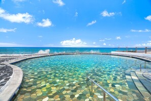 A clear pool with a hexagonal tile pattern, surrounded by a stone deck and a metal railing, overlooking a calm sea under a blue sky. - Oceanfront Complex - Updated Ground-floor Condo w/ AC & 2 Pools - Vrkcv (Kailua-Kona)