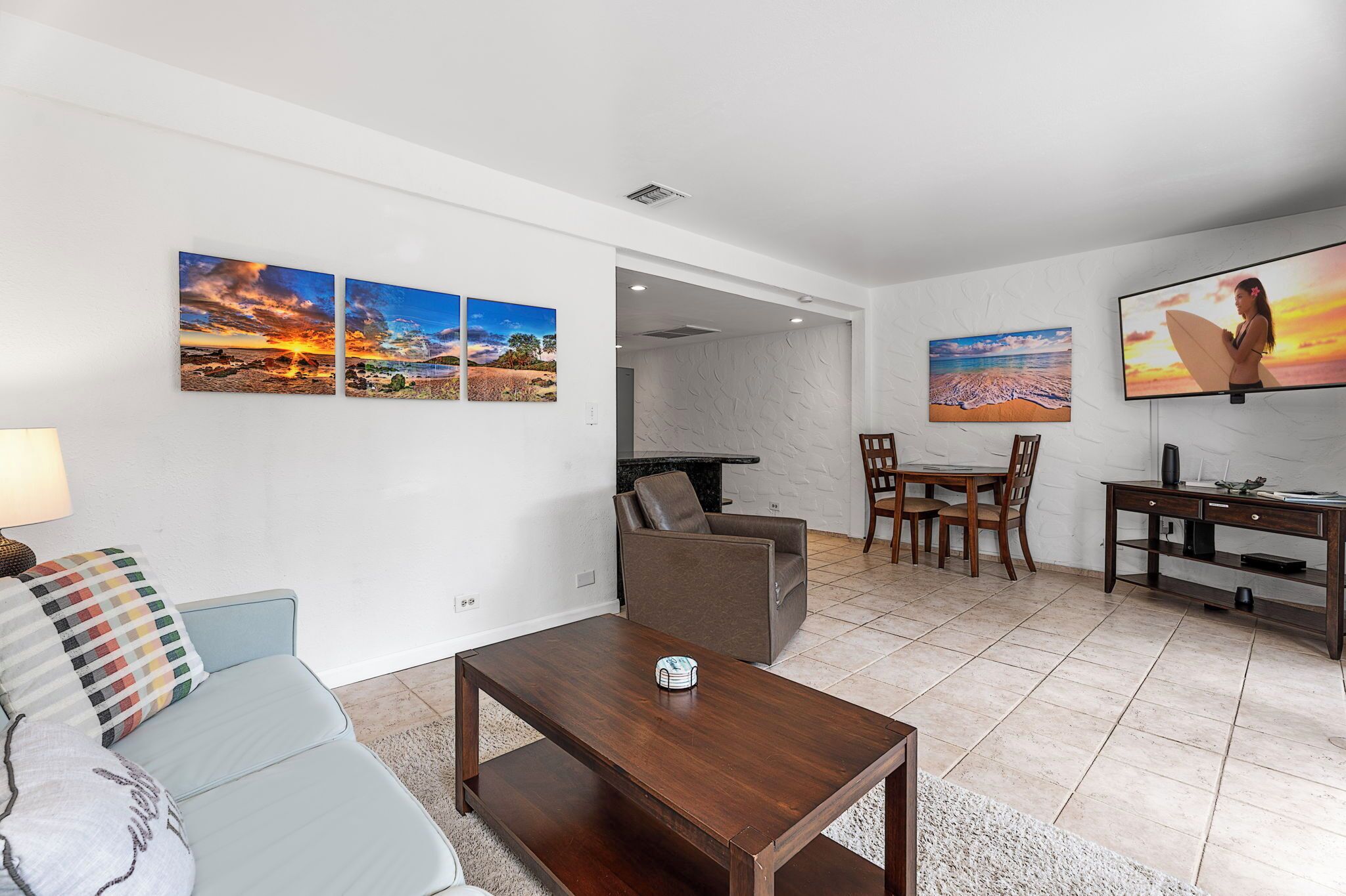 A modern living room with a sofa, coffee table, and a TV. There are two paintings on the wall and a dining area with chairs and a table.