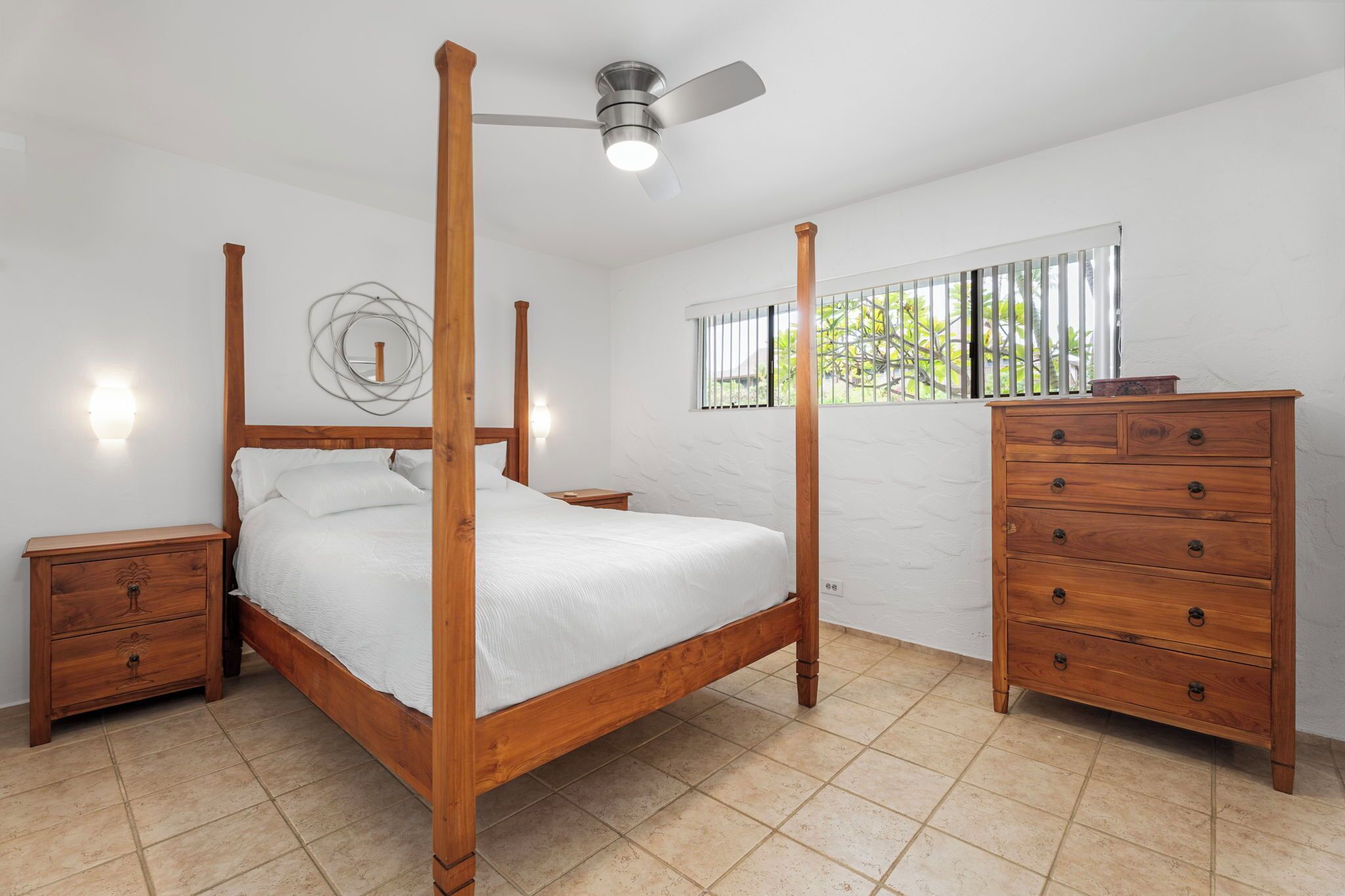 Une chambre à coucher avec un lit à baldaquin, des tables de chevet en bois, une commode, un ventilateur de plafond et une fenêtre avec des stores.