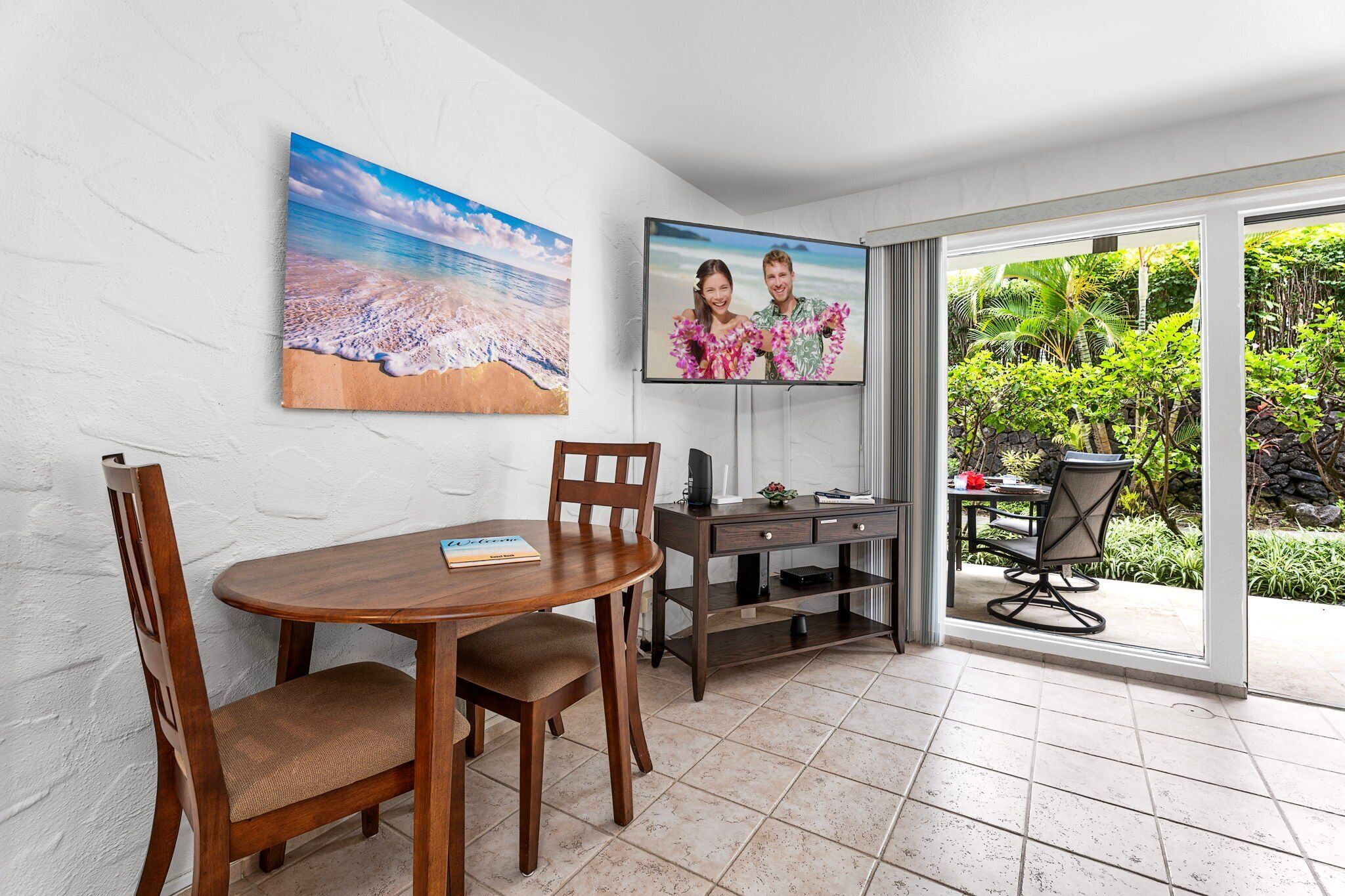 A dining area with a round table and chairs, a TV mounted on the wall, and a view of a tropical garden through a sliding glass door.