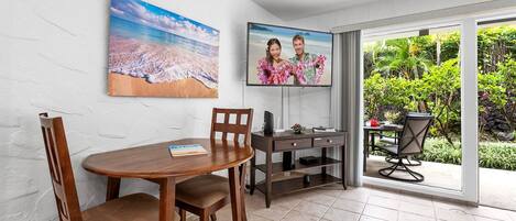 A dining area with a round table and chairs, a TV mounted on the wall, and a view of a tropical garden through a sliding glass door.