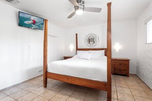 Ein Bett mit Himmel, ein hölzerner Rahmen, ein Deckenventilator, ein an der Wand montierter Fernseher der einen Surfer zeigt und ein kleiner Nachttisch mit einer Lampe.