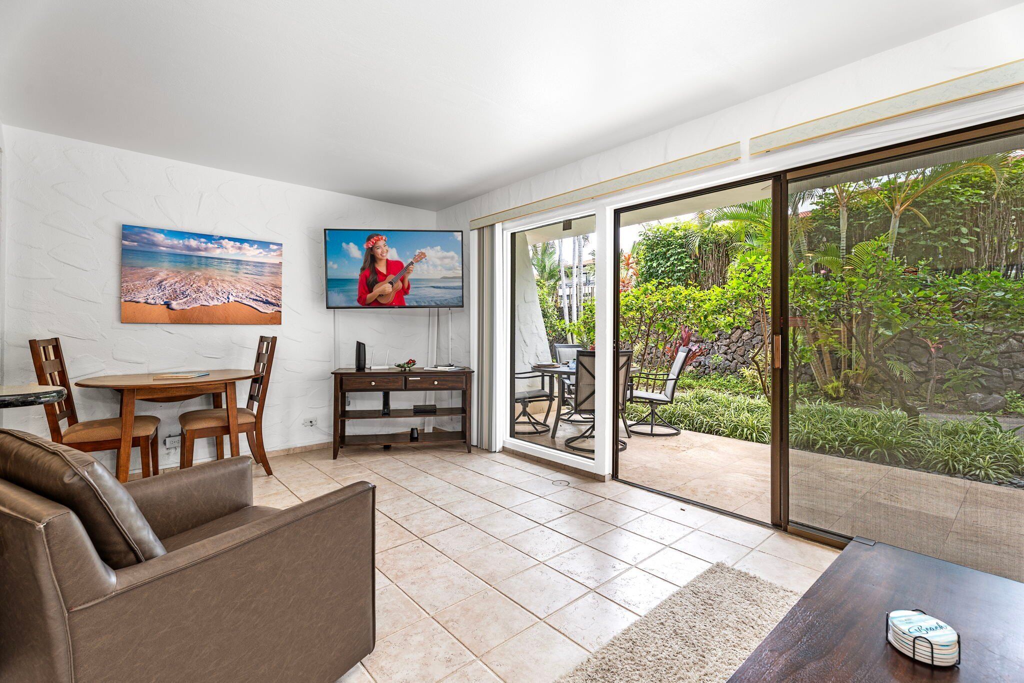 Una sala de estar con un sofá, una mesa de centro, un televisor y un comedor con sillas y una mesa. Hay una puerta corrediza de vidrio que da a un jardín con plantas y vistas al exterior.