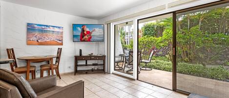 Una sala de estar con un sofá, una mesa de centro, un televisor y un comedor con sillas y una mesa. Hay una puerta corrediza de vidrio que da a un jardín con plantas y vistas al exterior.